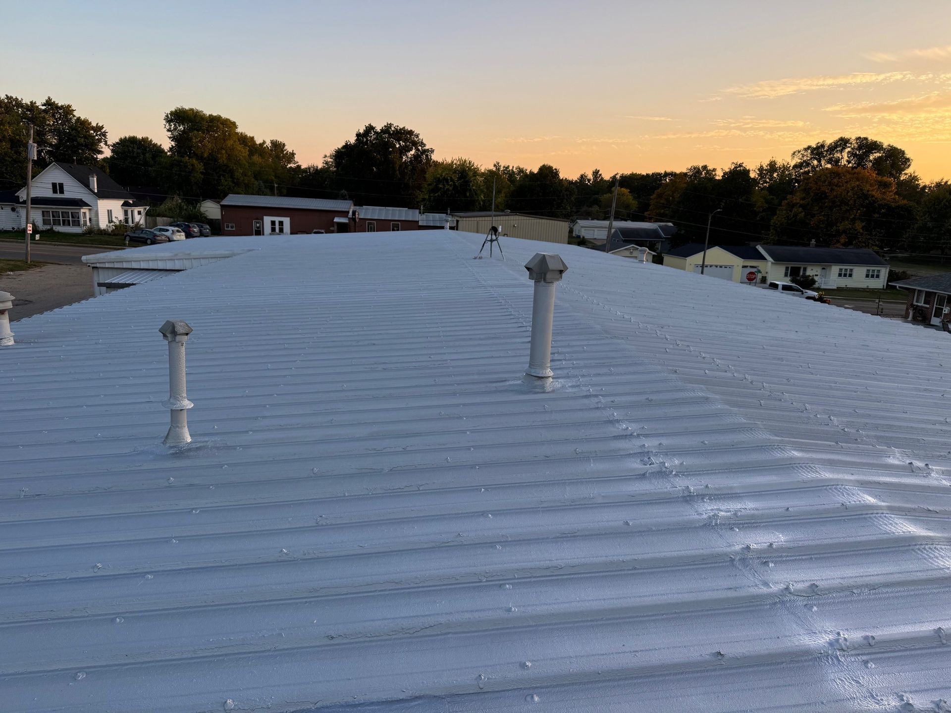 A person works on a large, freshly painted light-grey metal roof with two vent pipes at sunset.