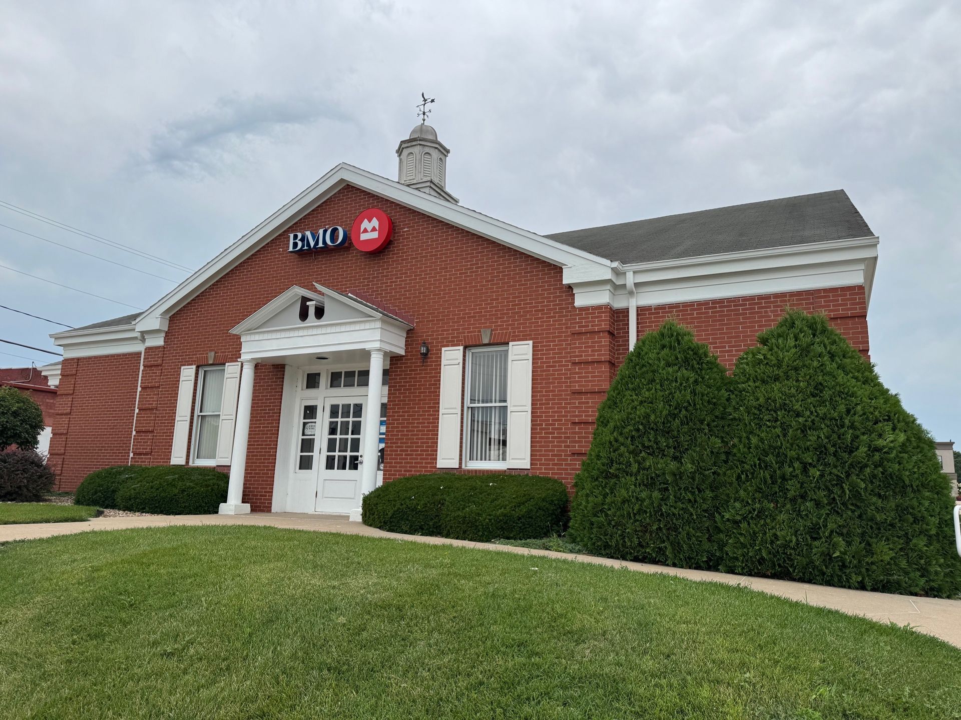 A red brick BMO bank branch with white trim, a cupola, and neatly trimmed green bushes on a sunny day.