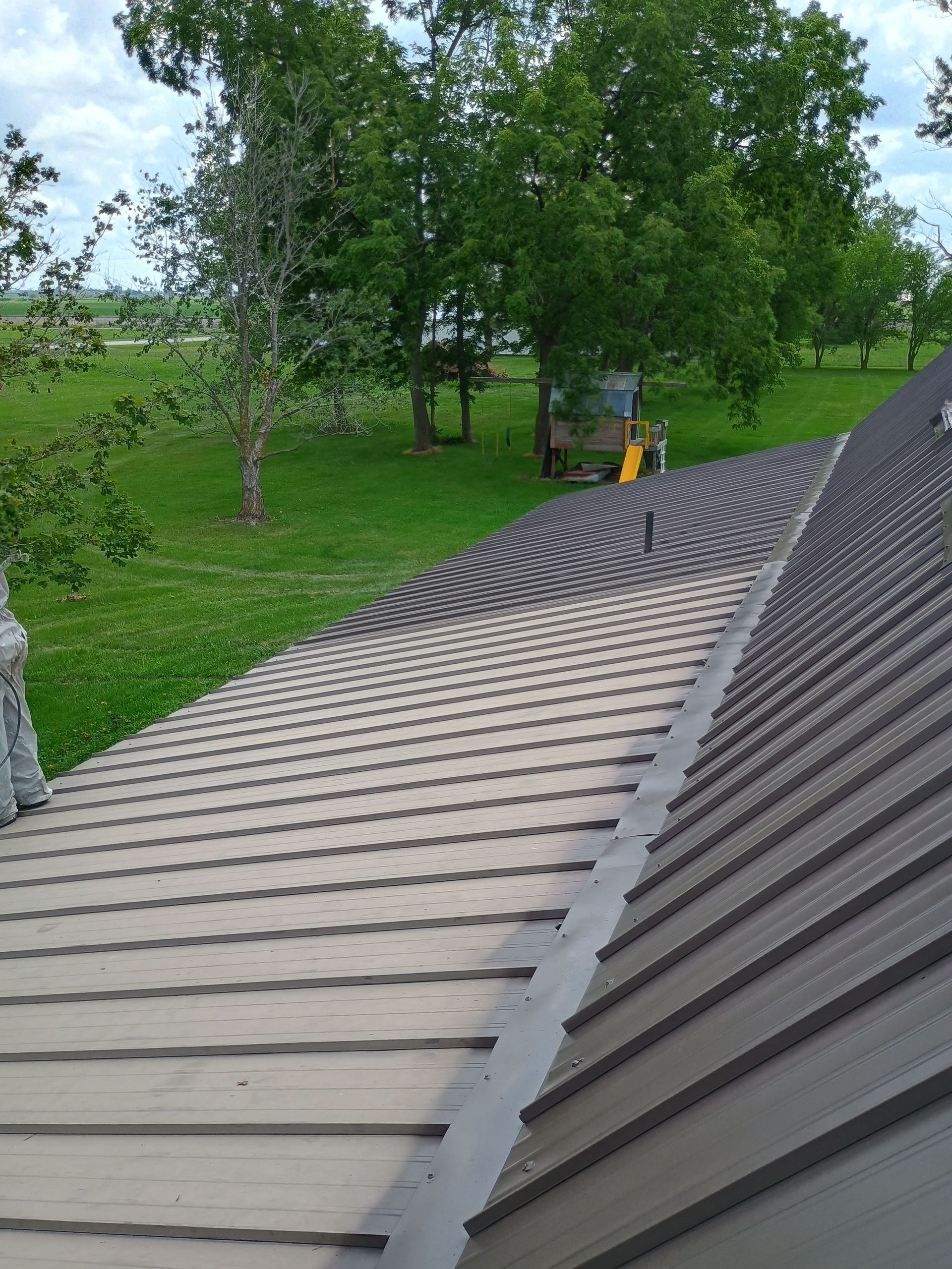 A view from a roof showing contrasting tan and dark grey metal roofing panels, with a green yard and trees in the background.