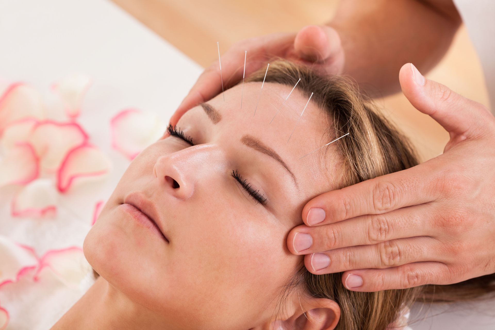 Hands gently massage a woman’s temple as she relaxes with eyes closed on a treatment bed.