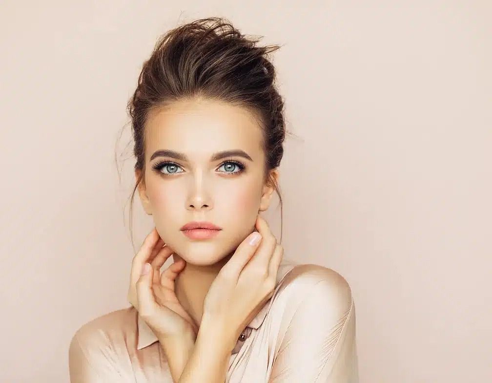 Portrait of a woman with hair in an updo, resting her hands on her neck against a soft beige background