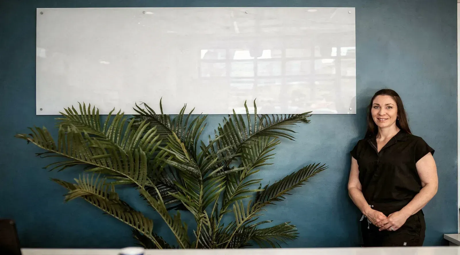 Woman standing beside a whiteboard and large potted plant in a classroom or office setting.