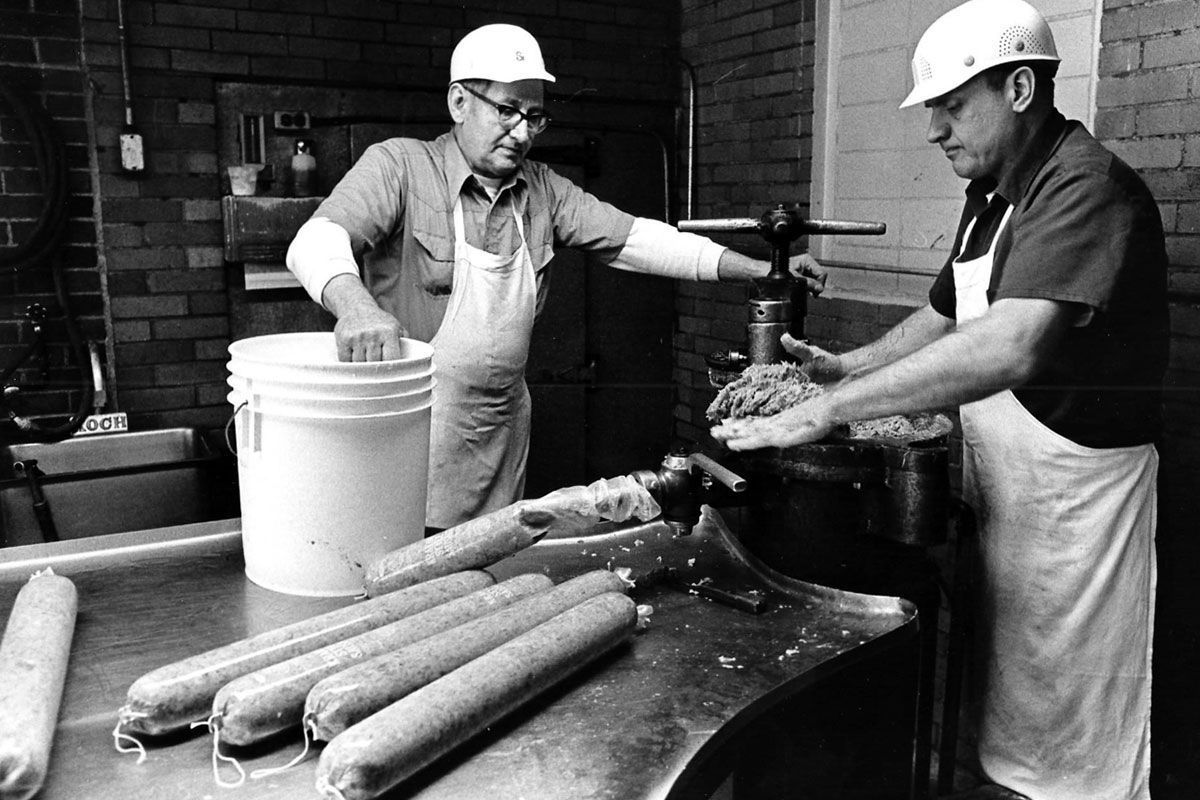 Two workers in hard hats prepare food at a counter, with buckets and rolls of dough nearby.