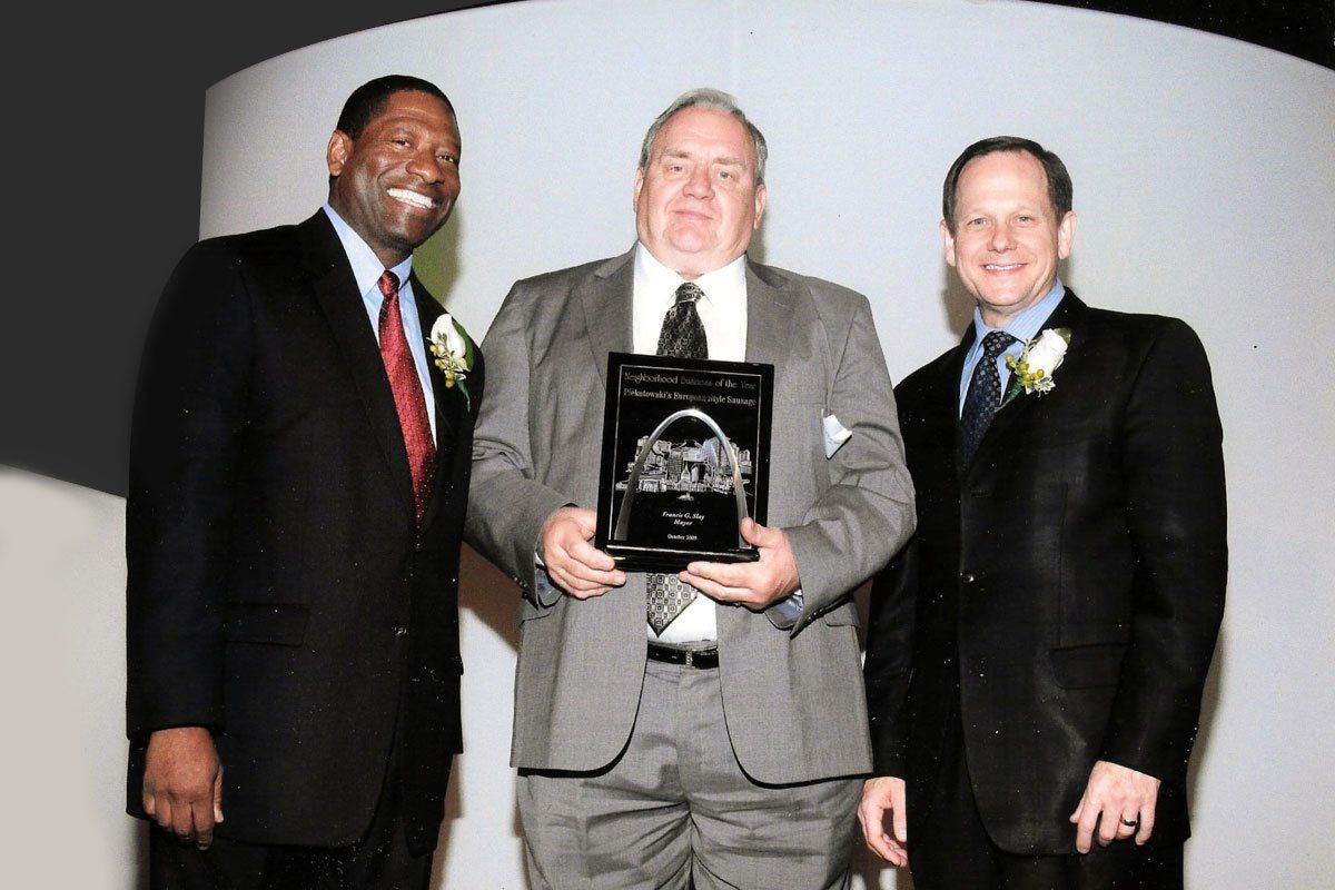 Three men in suits posing at an awards ceremony, one holding a plaque.