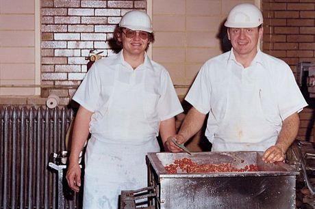 Two workers in white uniforms and hard hats stand by a metal processing machine indoors.