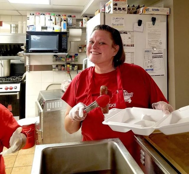 Volunteer in red shirt serving food at a restaurant counter, smiling while holding a takeout container.