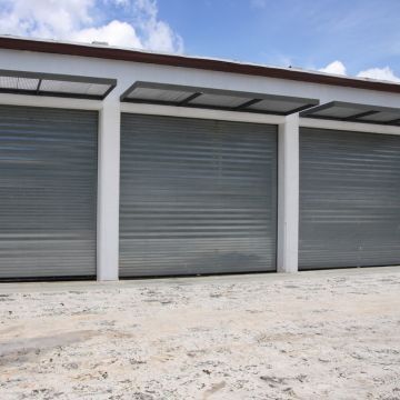 Three closed gray roll-up garage doors on a white building, set on a concrete surface, under a partly cloudy sky.