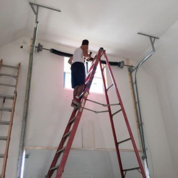 Person on a ladder, installing garage door hardware. Ceiling is white, and the ladder is red.