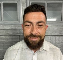 Man smiling, wearing a suit jacket and earbuds outdoors.