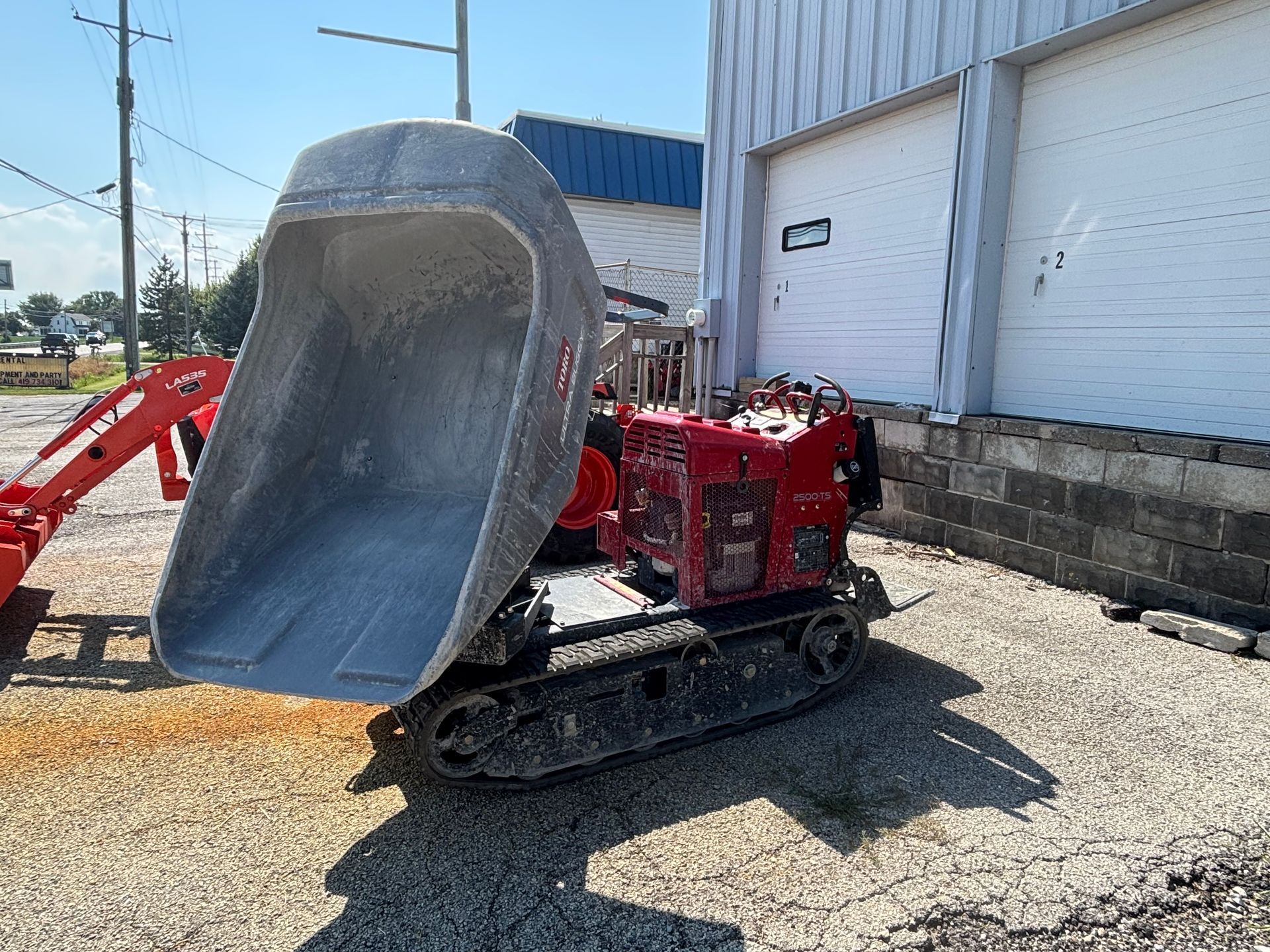 Red track dumper with raised gray bucket, next to a building.