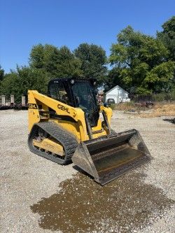 Yellow Gehl track loader with bucket parked on gravel. Blue sky and trees in background.