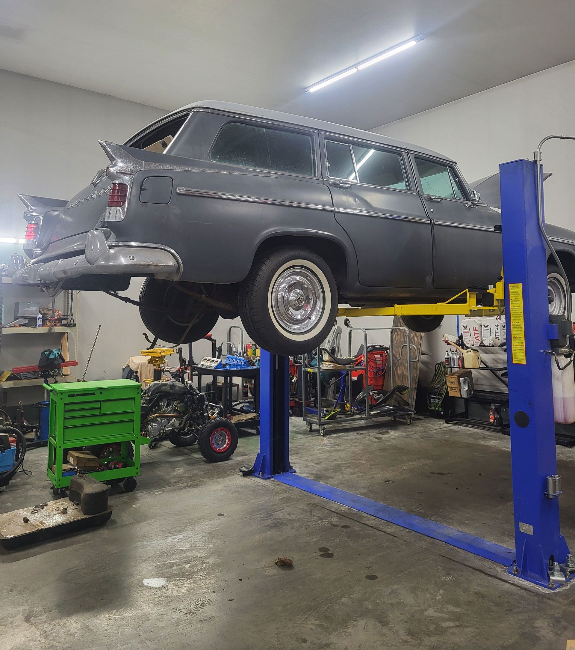 A gray station wagon undergoing restoration, elevated on a blue two-post lift inside an auto repair shop.