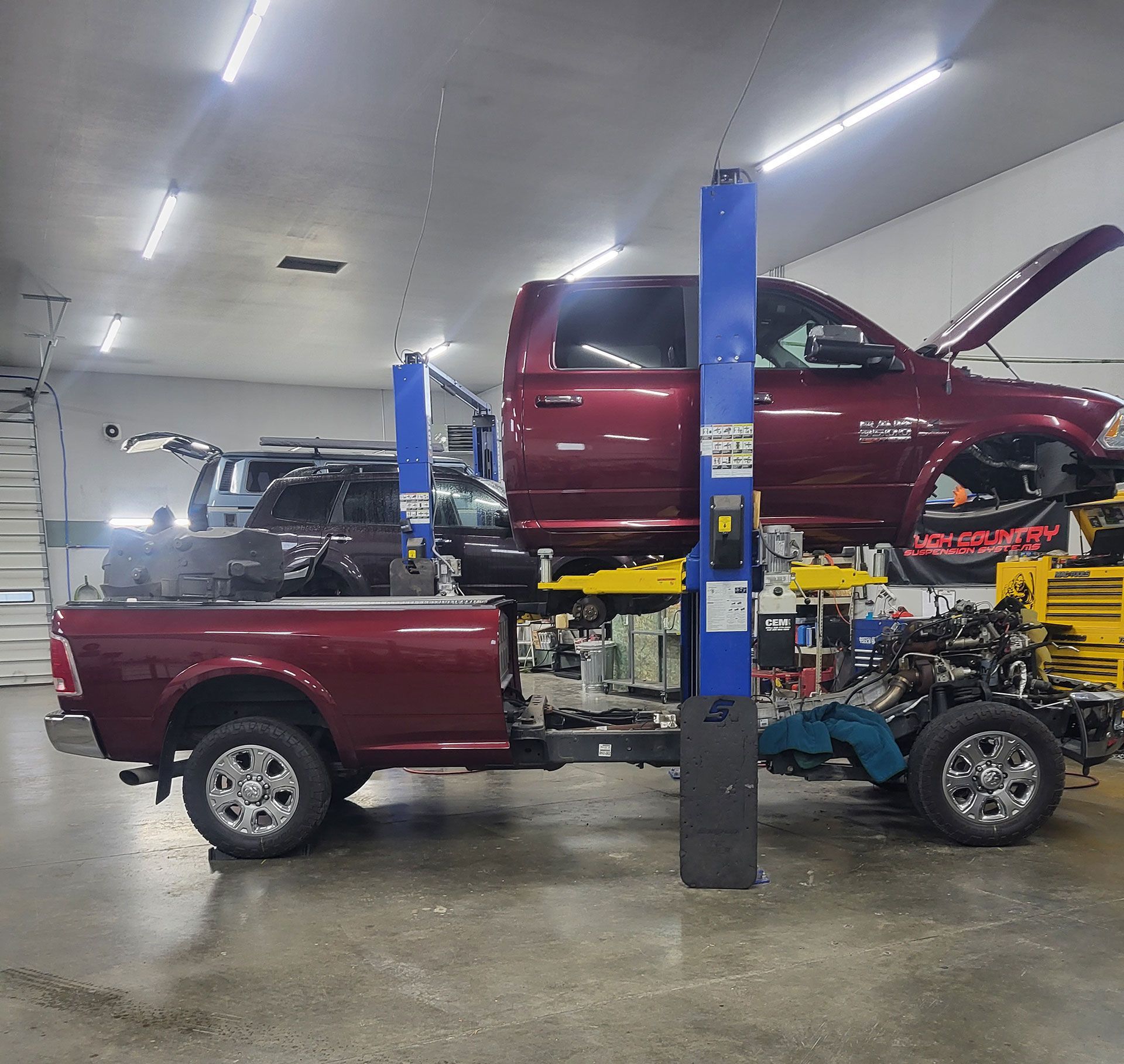 A maroon pickup truck with its cab lifted on a hydraulic auto lift in a brightly lit repair shop.