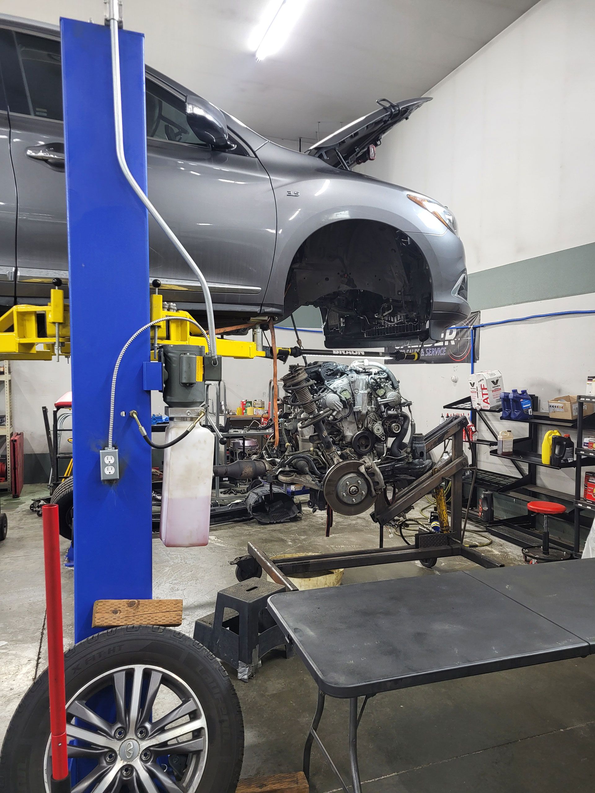 A gray car raised on a hydraulic lift in a garage, with an engine detached and resting on a stand underneath.