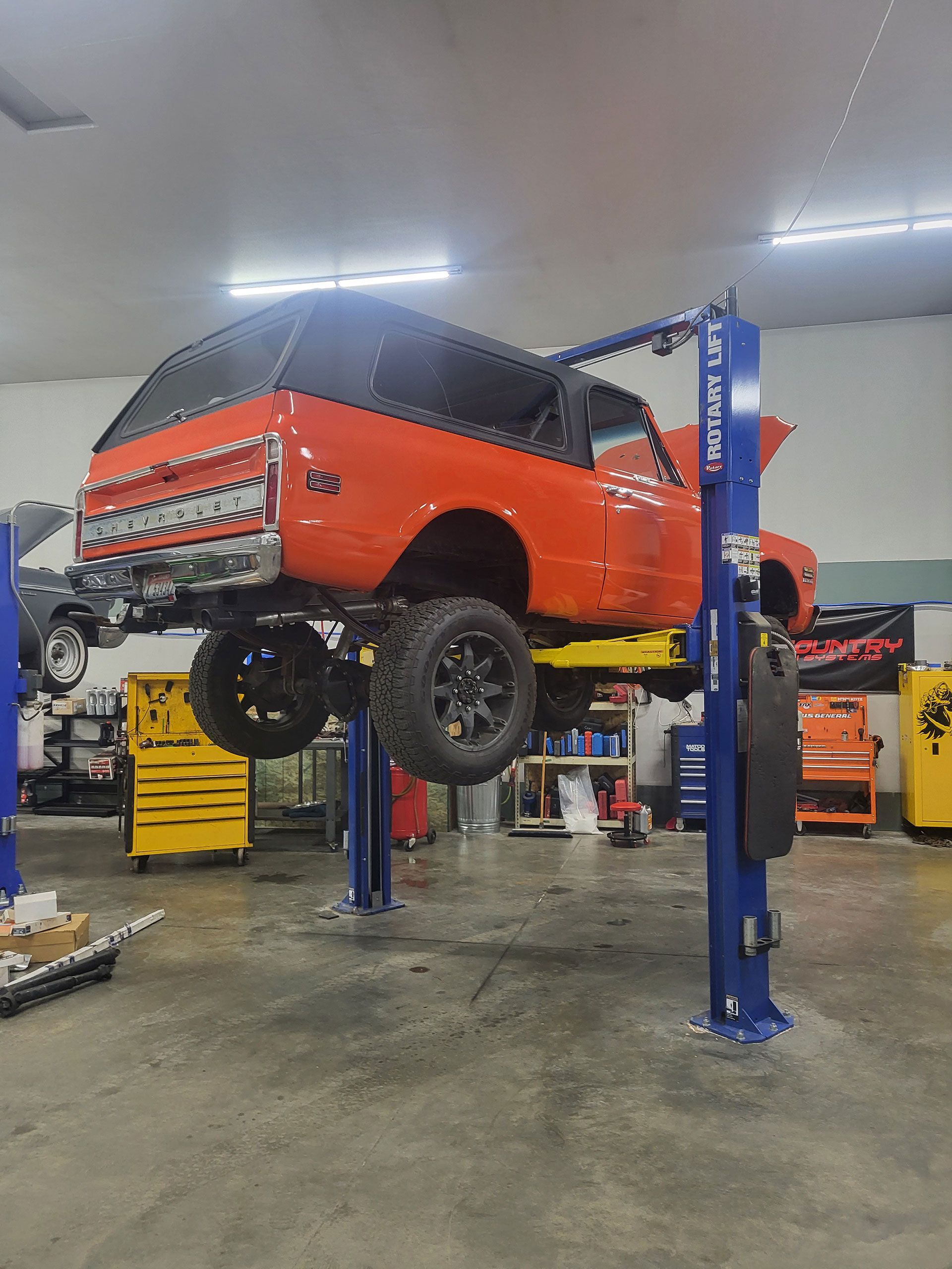 An orange vintage Ford Bronco elevated on a blue two-post hydraulic vehicle lift in an automotive repair shop.