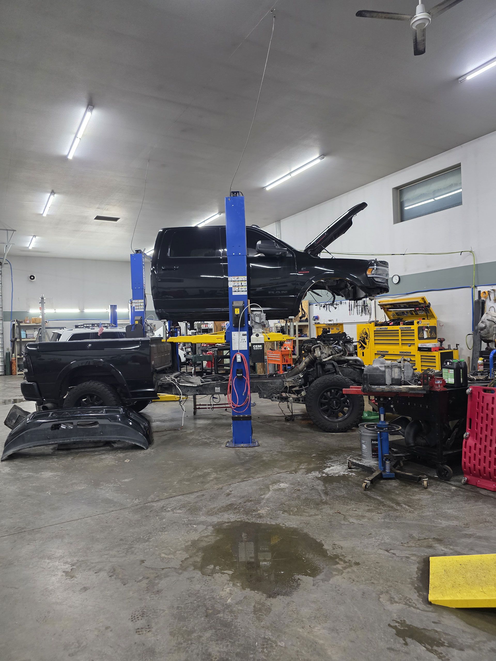 A black truck is lifted on a blue hydraulic lift inside an automotive repair shop with tools and equipment nearby.