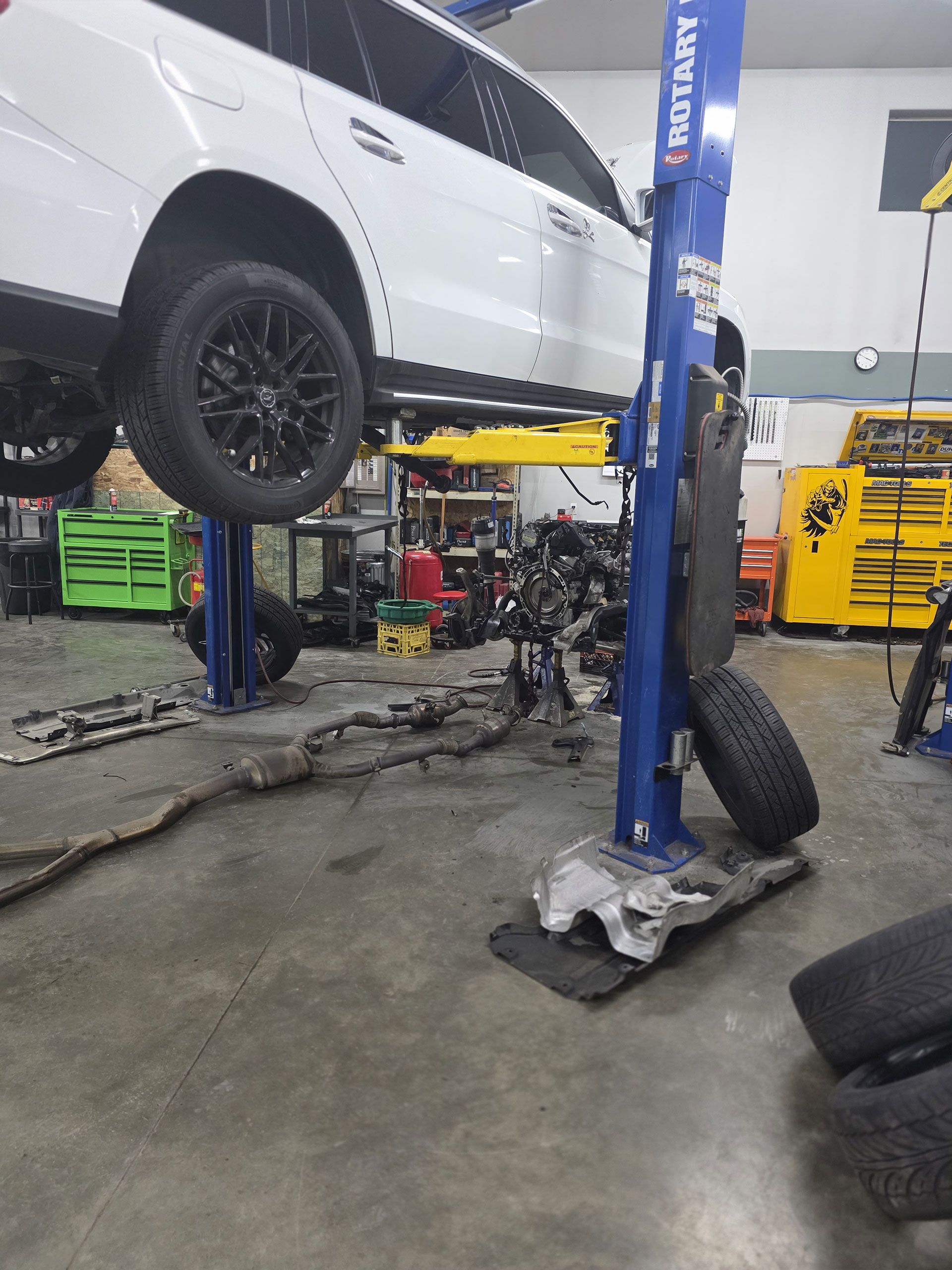 A white SUV elevated on a blue automotive lift in a workshop, with car parts and tires on the floor.