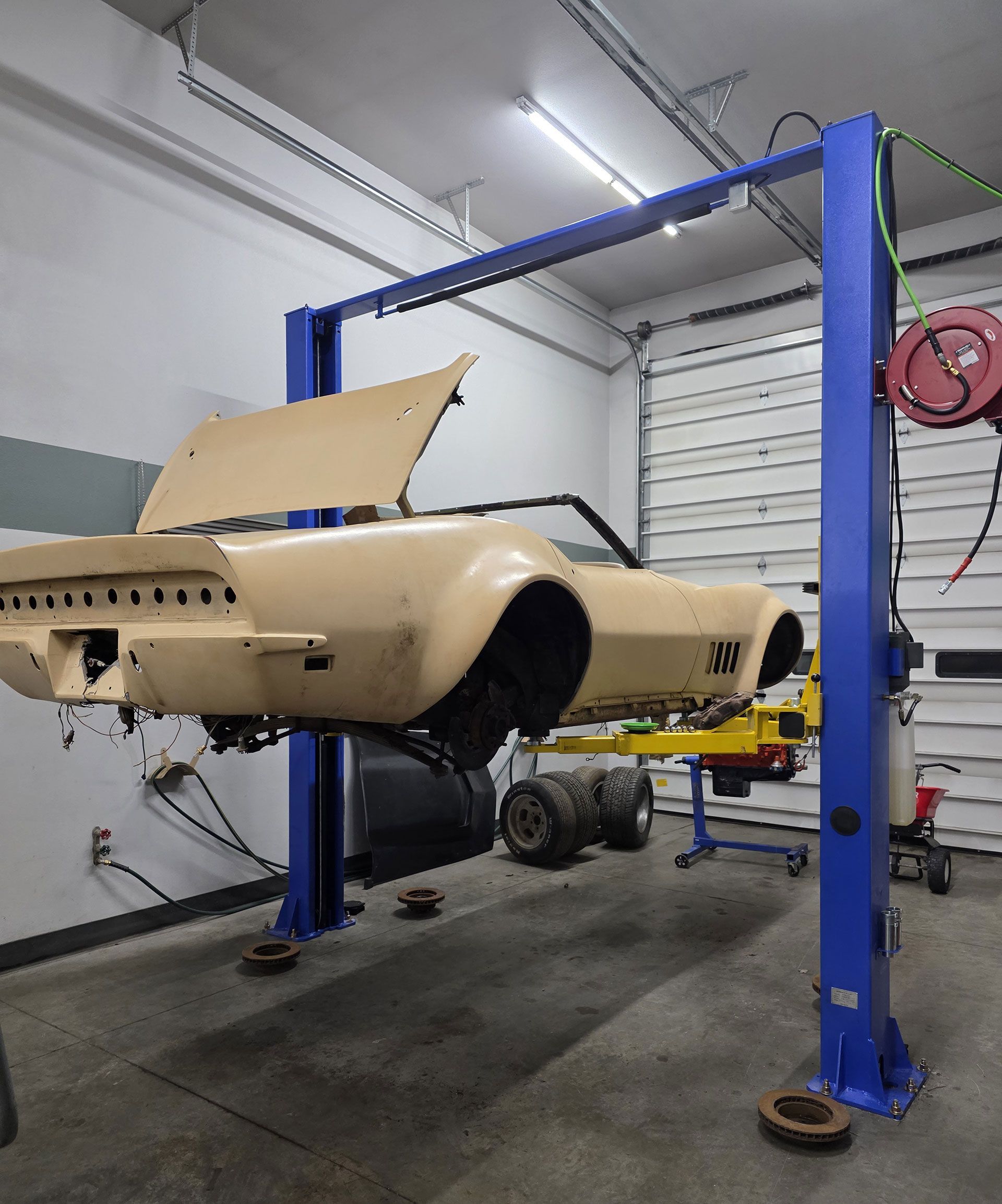 A cream-colored vintage convertible car body undergoing restoration, elevated on a blue hydraulic lift in a garage.