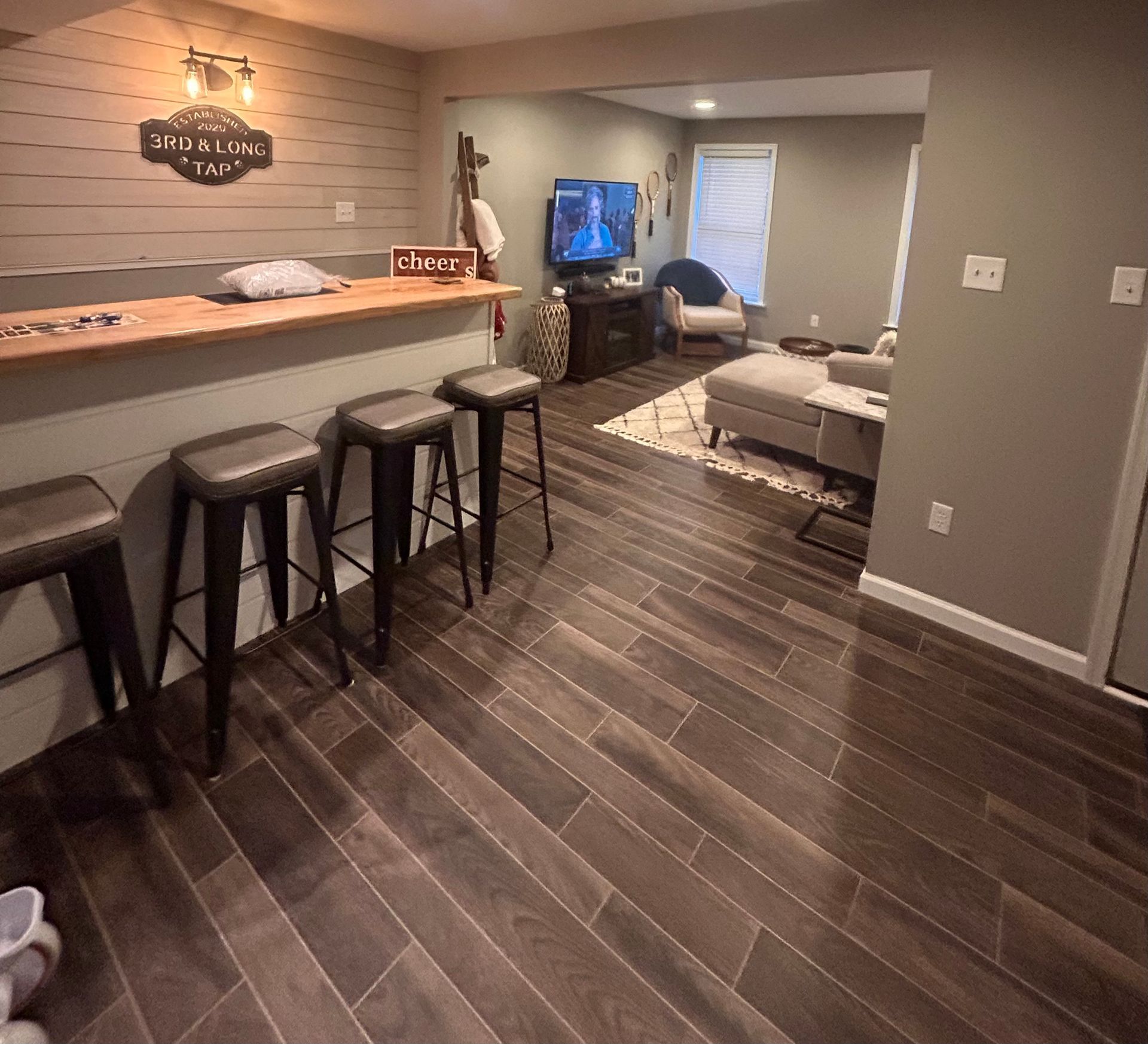 Basement lounge with wood bar, stools, TV, sectional sofa, and patterned rug.