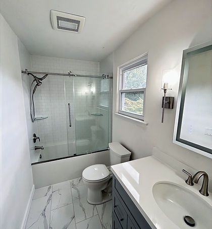 Bright white modern bathroom with glass shower, toilet, vanity sink, and marble-look floor.