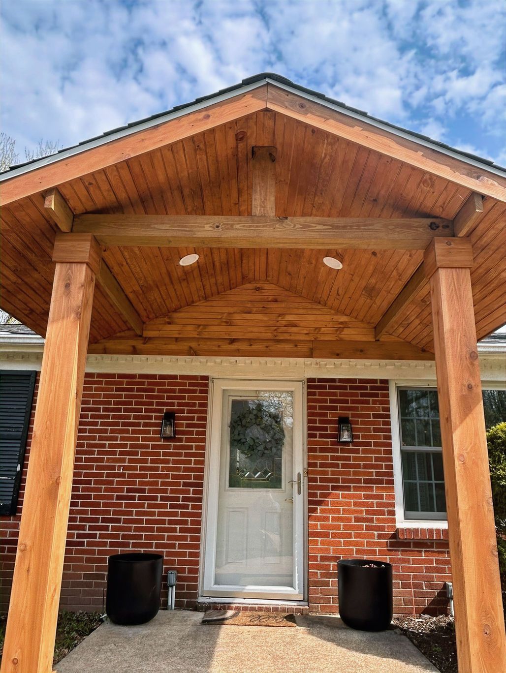 Brick house front porch with wooden roof, white door, and two black planters.