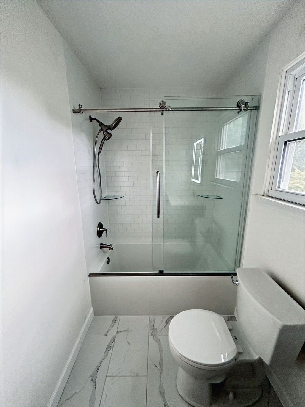 Small white bathroom with a glass shower, toilet, and window on the right.