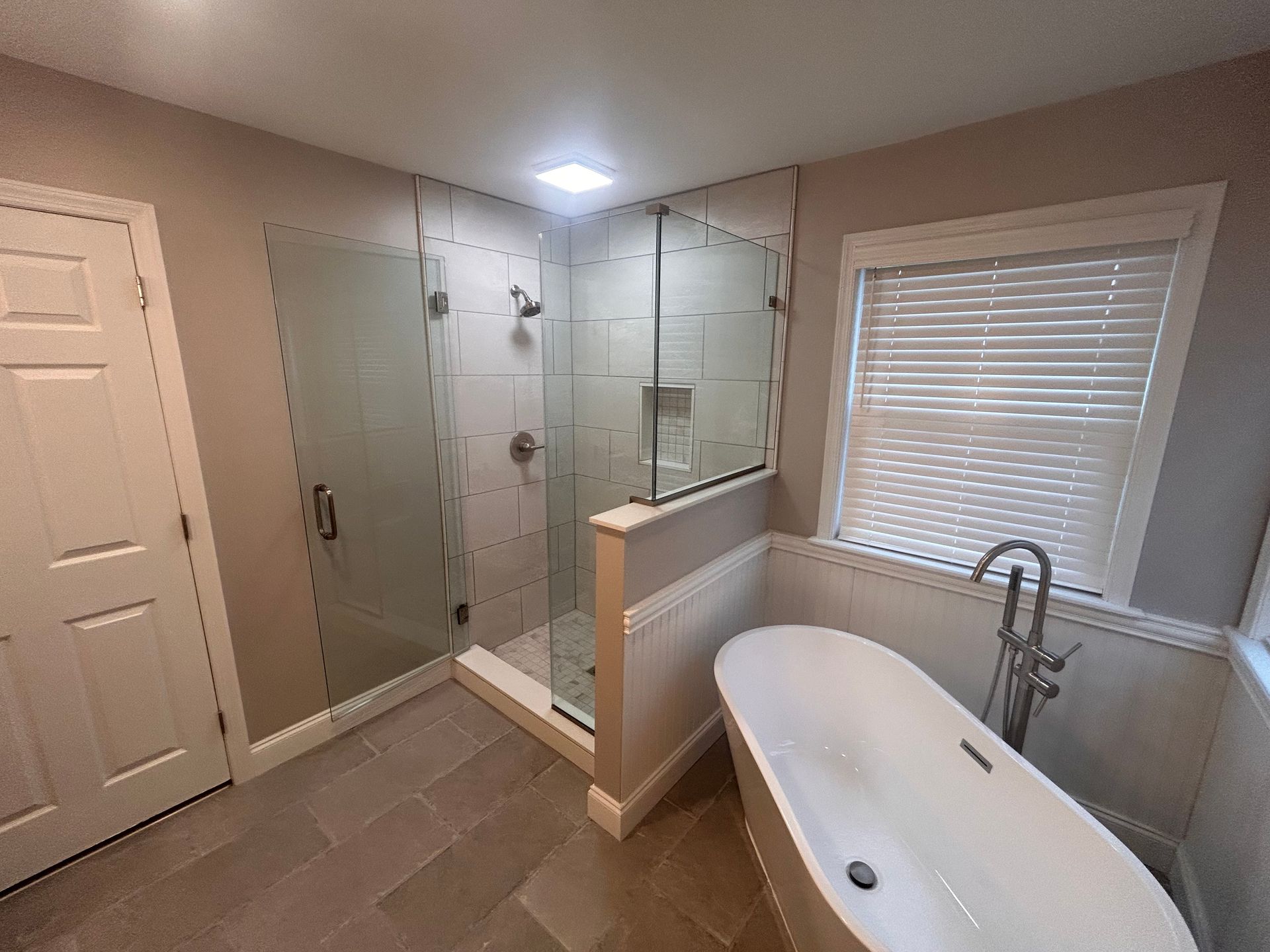 Modern bathroom with glass shower, freestanding tub, window blinds, and gray tile floor