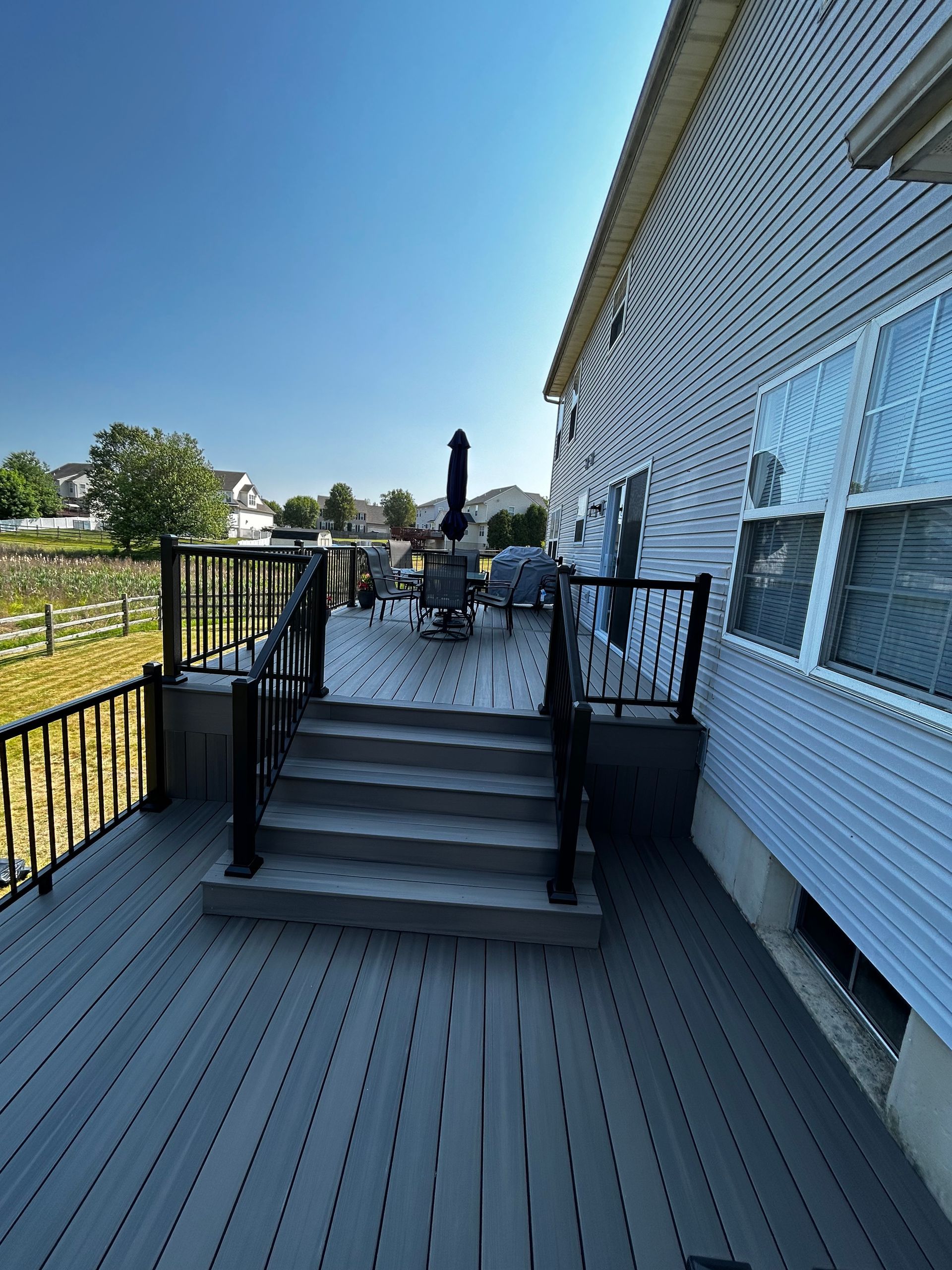 Back deck stairs leading to a patio with chairs, beside a white house with blue patterned siding