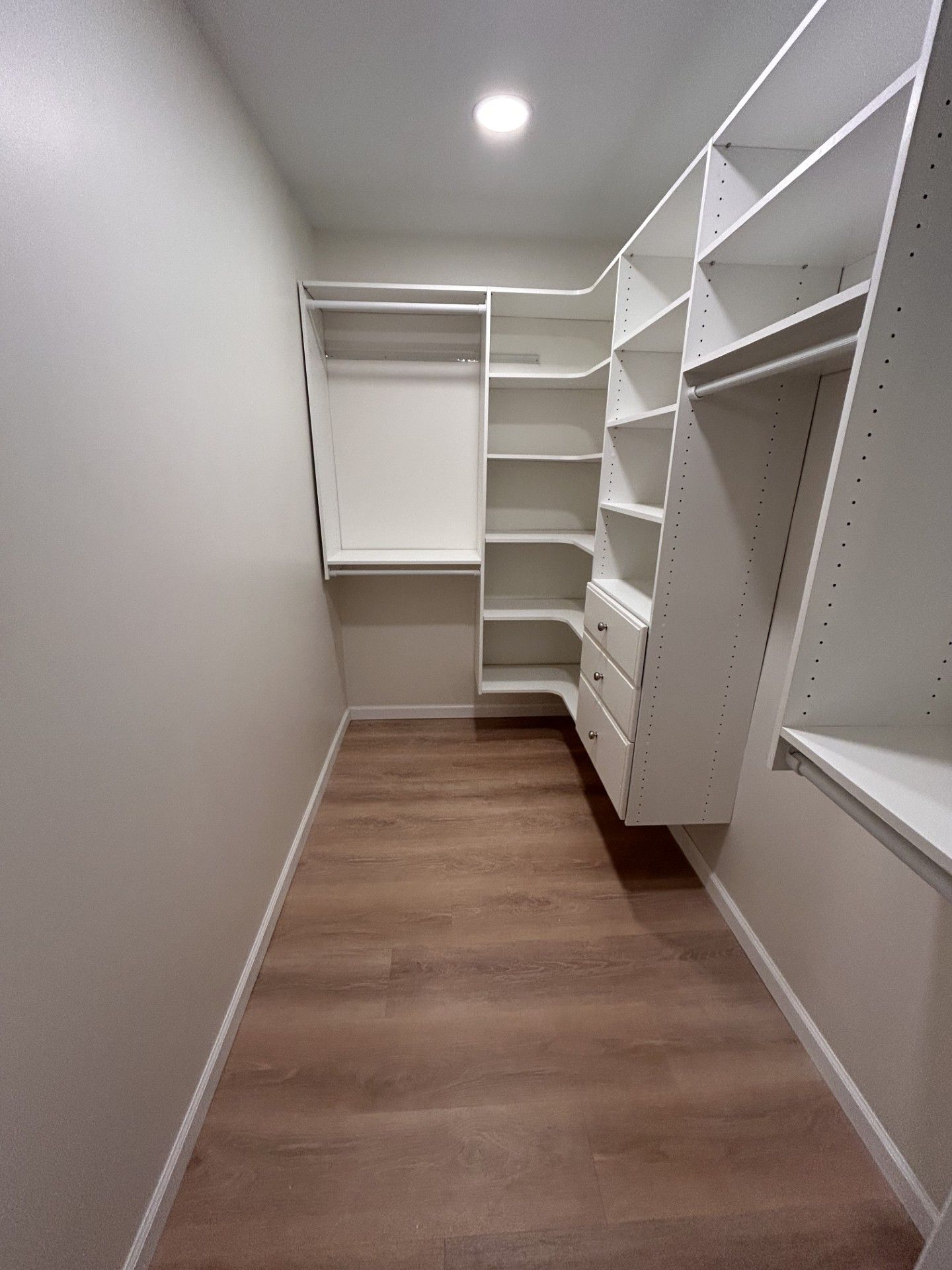 Empty walk-in closet with white shelves and drawers, wood floor, and a recessed ceiling light.