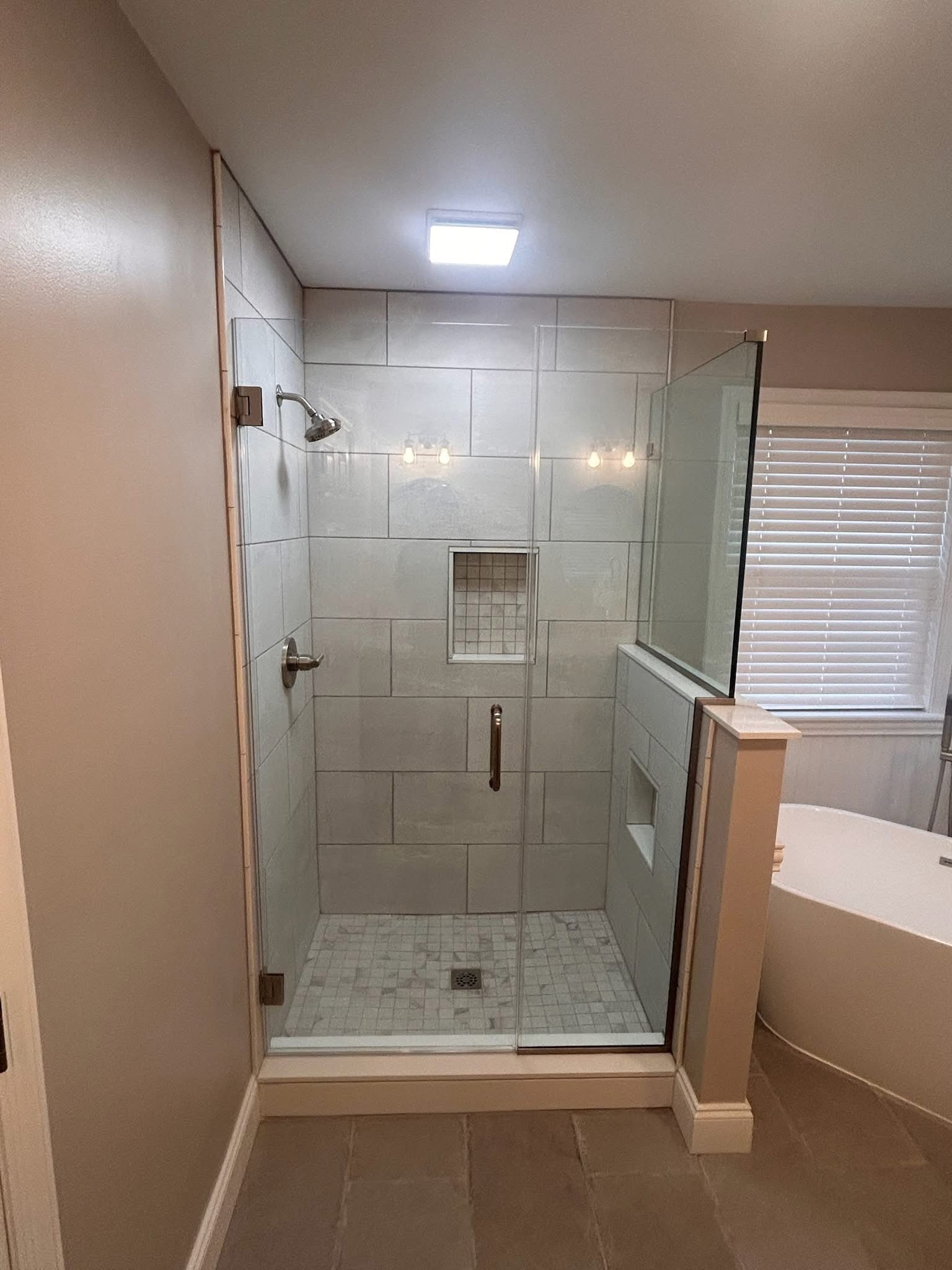 Bathroom with glass-enclosed shower, white tile walls, and a window with blinds nearby.
