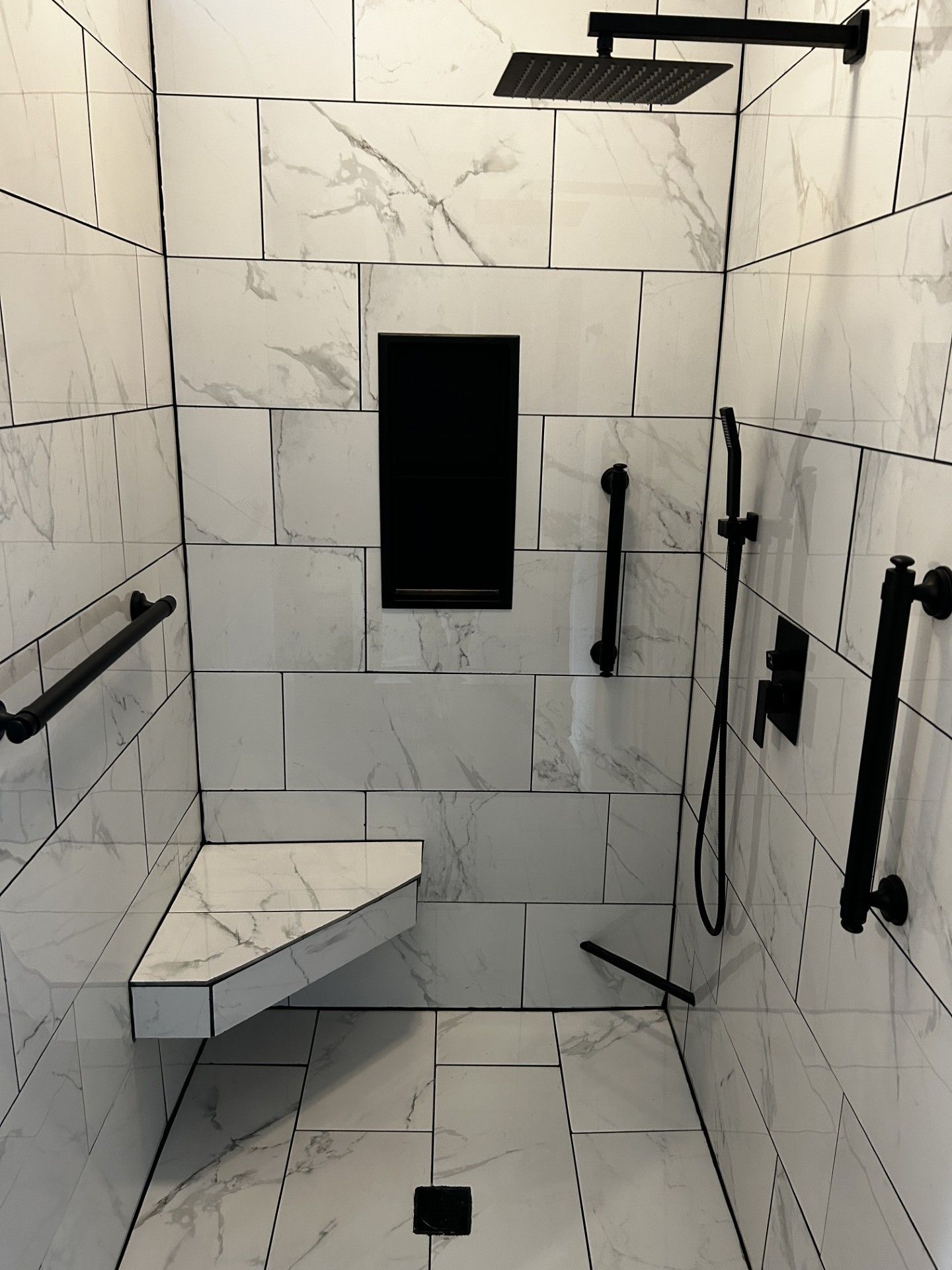Shower with white tile, black fixtures, bench, grab bars, and handheld showerhead.