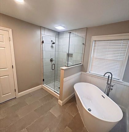 Modern bathroom with glass shower, freestanding tub, gray tile, and window blinds