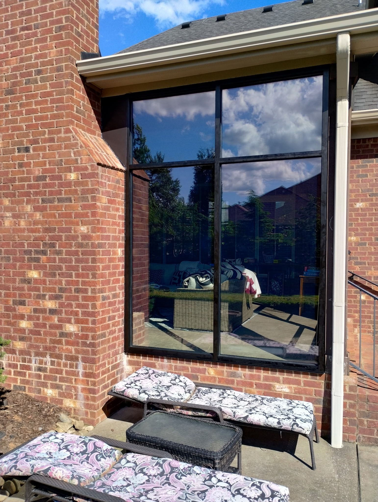 A brick house exterior with a large, dark-tinted window reflecting the sky and two outdoor lounge chairs on a patio. 