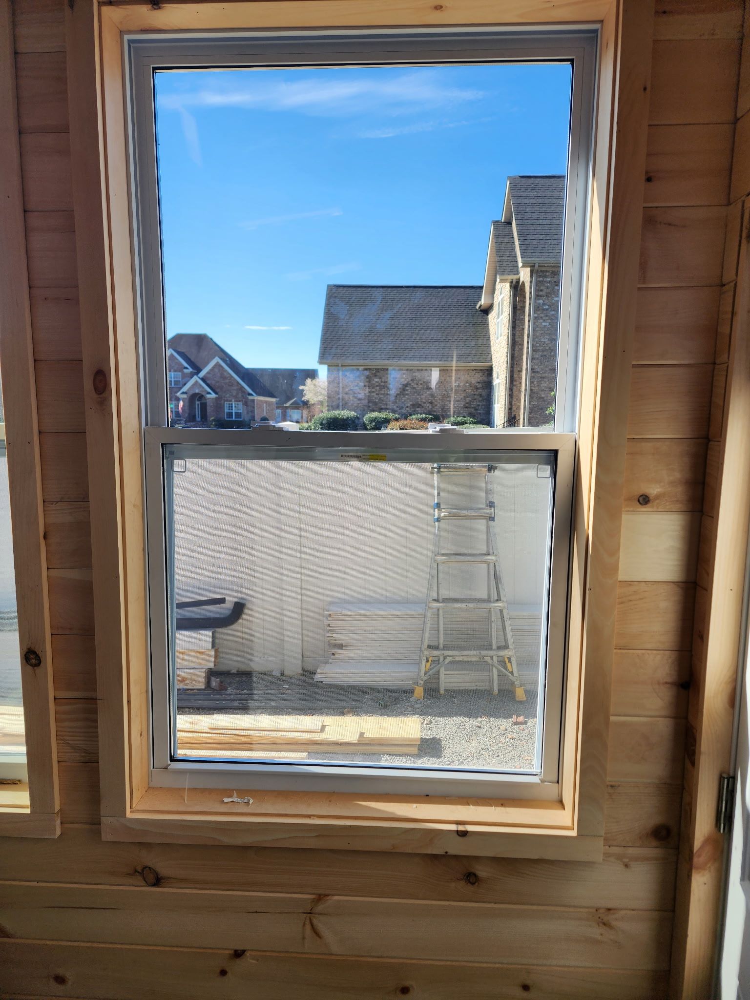 A single-hung window with a screen is framed by light-colored wood walls, looking out at a residential outdoor scene. 