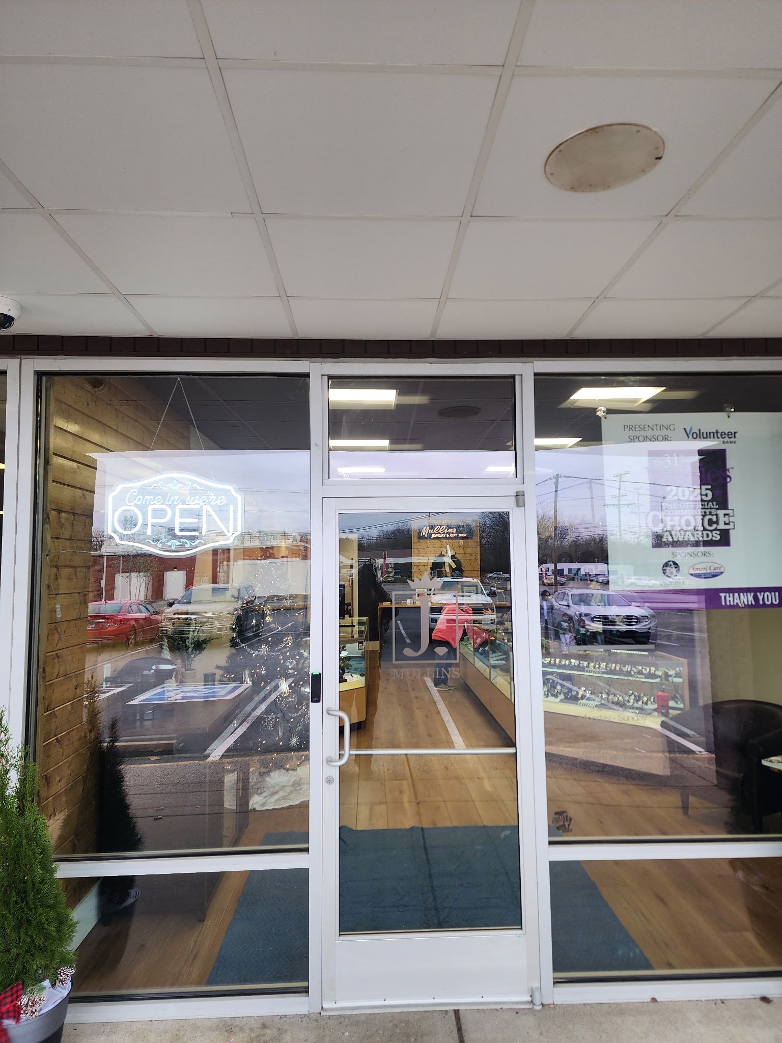 The exterior entrance of a retail store with a glass door, a neon open sign, and a view into the bright, wood-floored shop. 