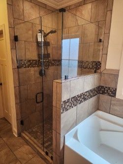 A corner shower with tan stone tile, a glass door, and a decorative mosaic border next to a white bathtub. 