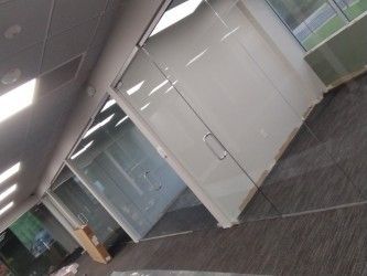 Modern office interior featuring glass-paneled walls, metal door handles, recessed ceiling lights, and grey carpeting. 