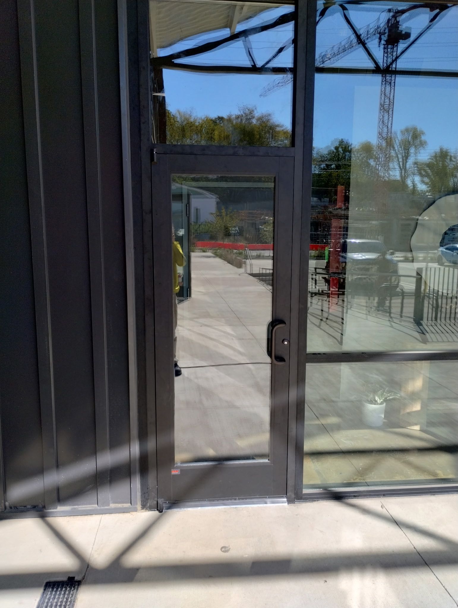 A dark-framed glass door with a push handle installed in a metal-paneled building exterior facing a concrete walkway. 