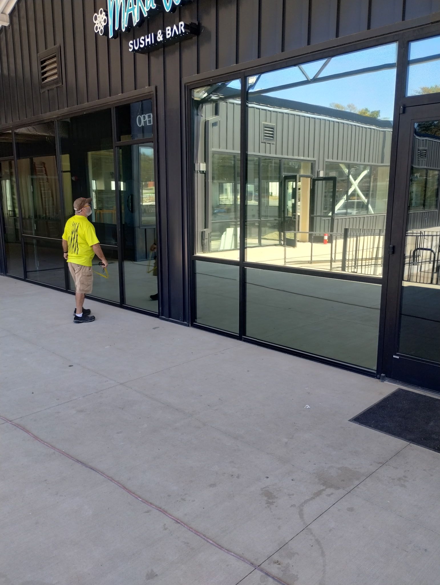 A person in a bright yellow shirt stands outside a storefront with a glass front and dark metal siding. 