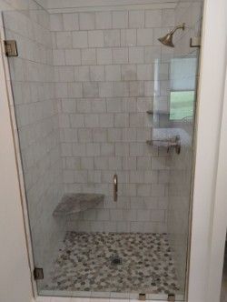 A walk-in shower with white tiled walls, a pebble floor, a built-in corner seat, and a glass door. 