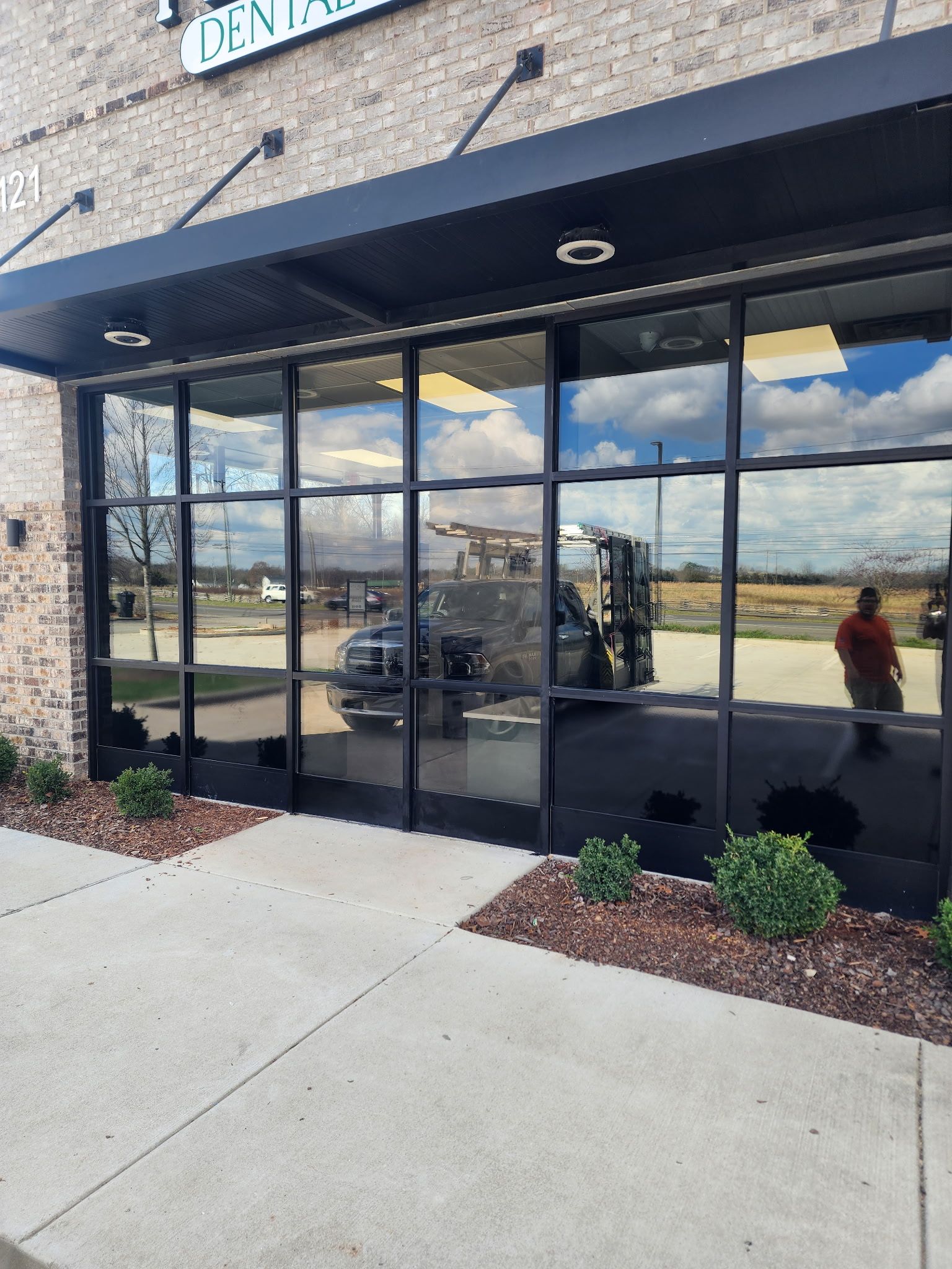 Exterior view of a modern dental office storefront with large, dark tinted windows reflecting a truck in the parking lot. 
