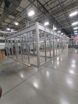 A metal-framed glass office enclosure under construction inside a large warehouse facility with overhead lighting. 