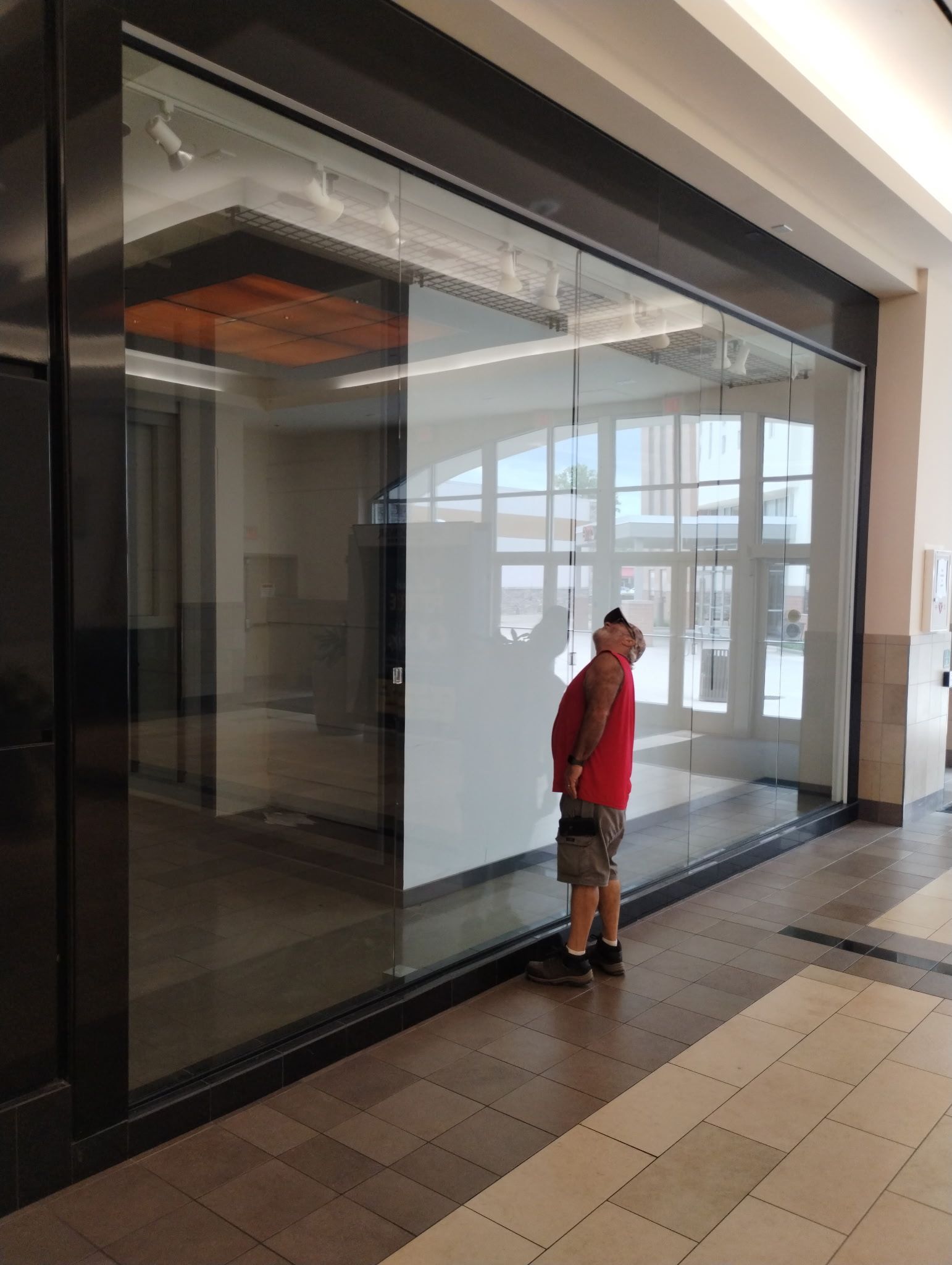 A person in a red shirt and shorts stands inside a mall corridor looking up at a large glass storefront window. 