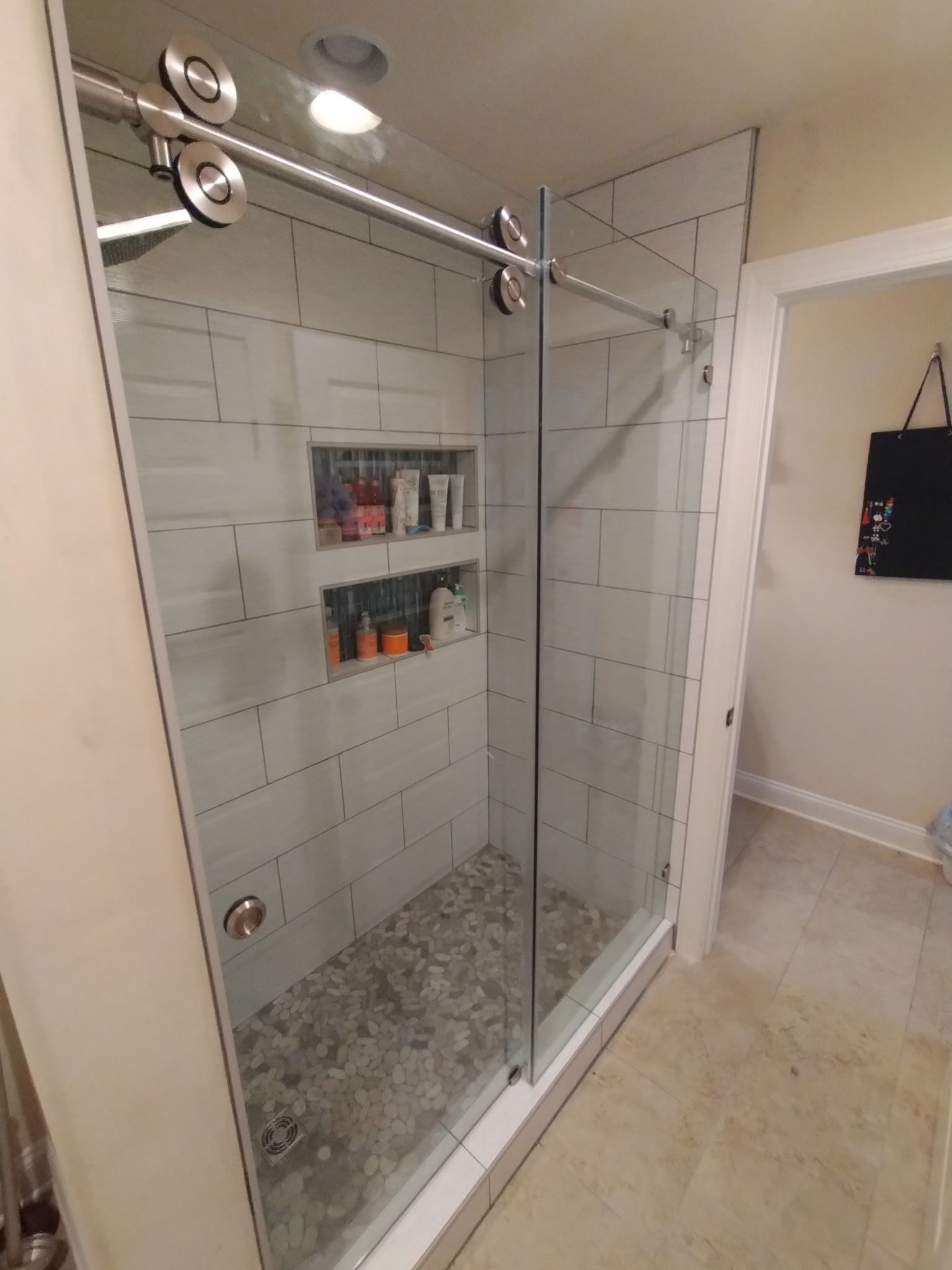 A tiled walk-in shower with a sliding glass door, overhead lighting, and two recessed shelving niches filled with bottles. 