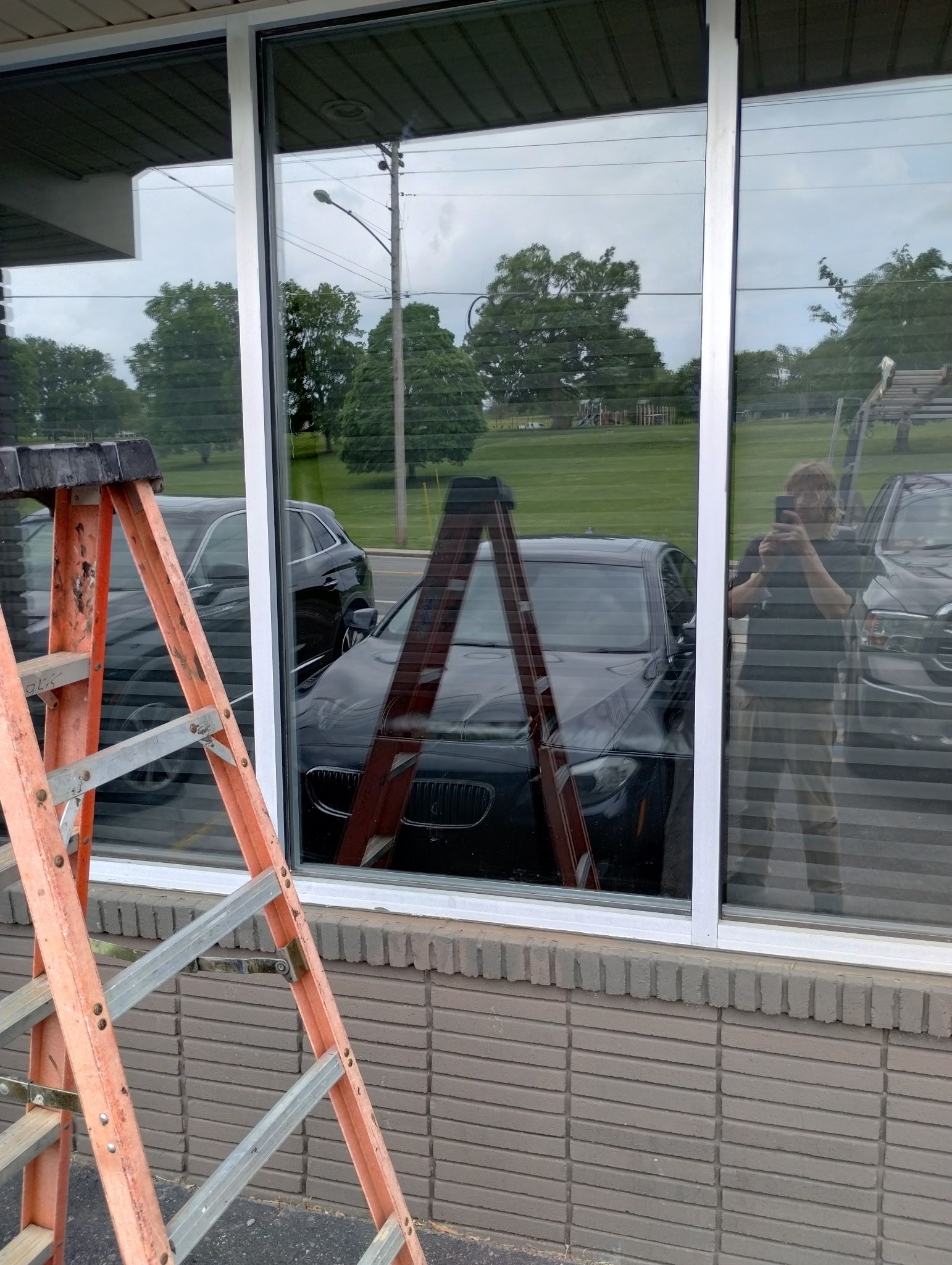 A person stands outside holding a camera, reflected in large windows along with a step ladder and cars in a parking lot. 