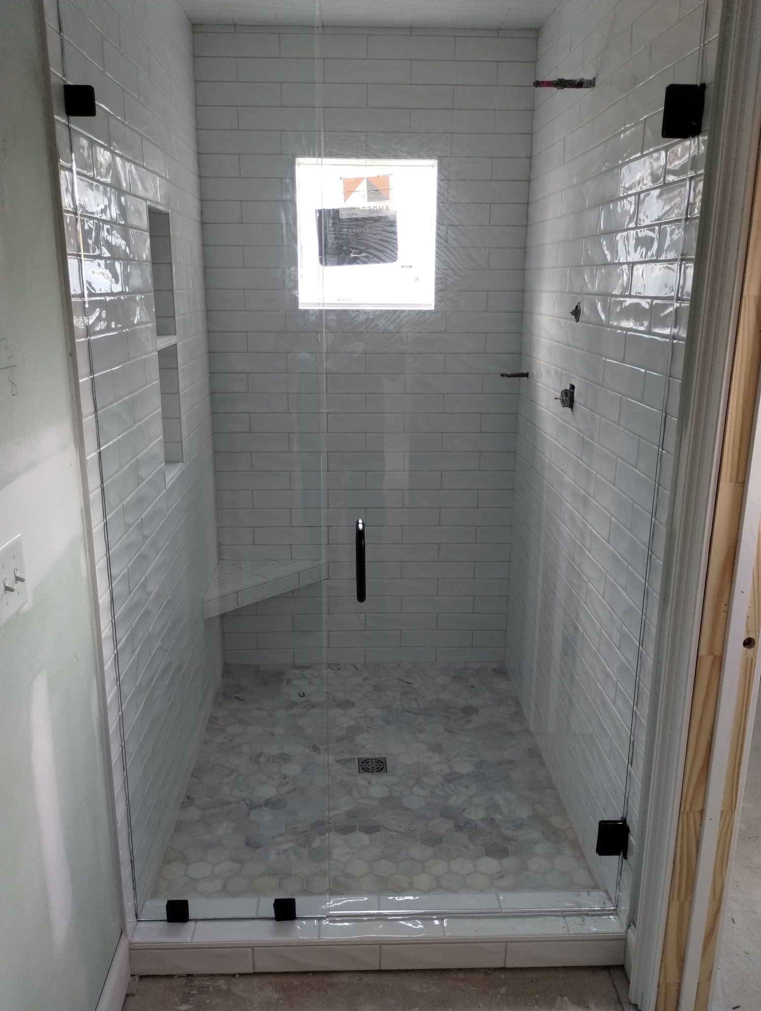 A tiled walk-in shower with a glass door, a built-in bench, a small window, and grey stone floor tiling. 