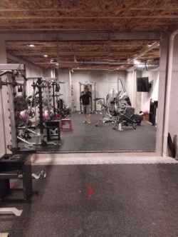 A person stands in a home gym with fitness equipment, including a squat rack and exercise bike, reflected in a large mirror. 