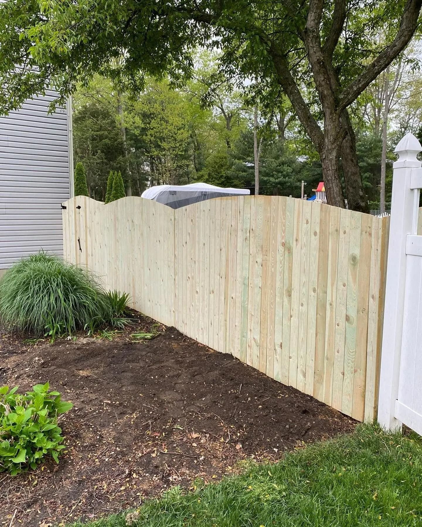 Tall wooden fence beside a garden bed and grass, with trees overhead and a white gate post on the right.