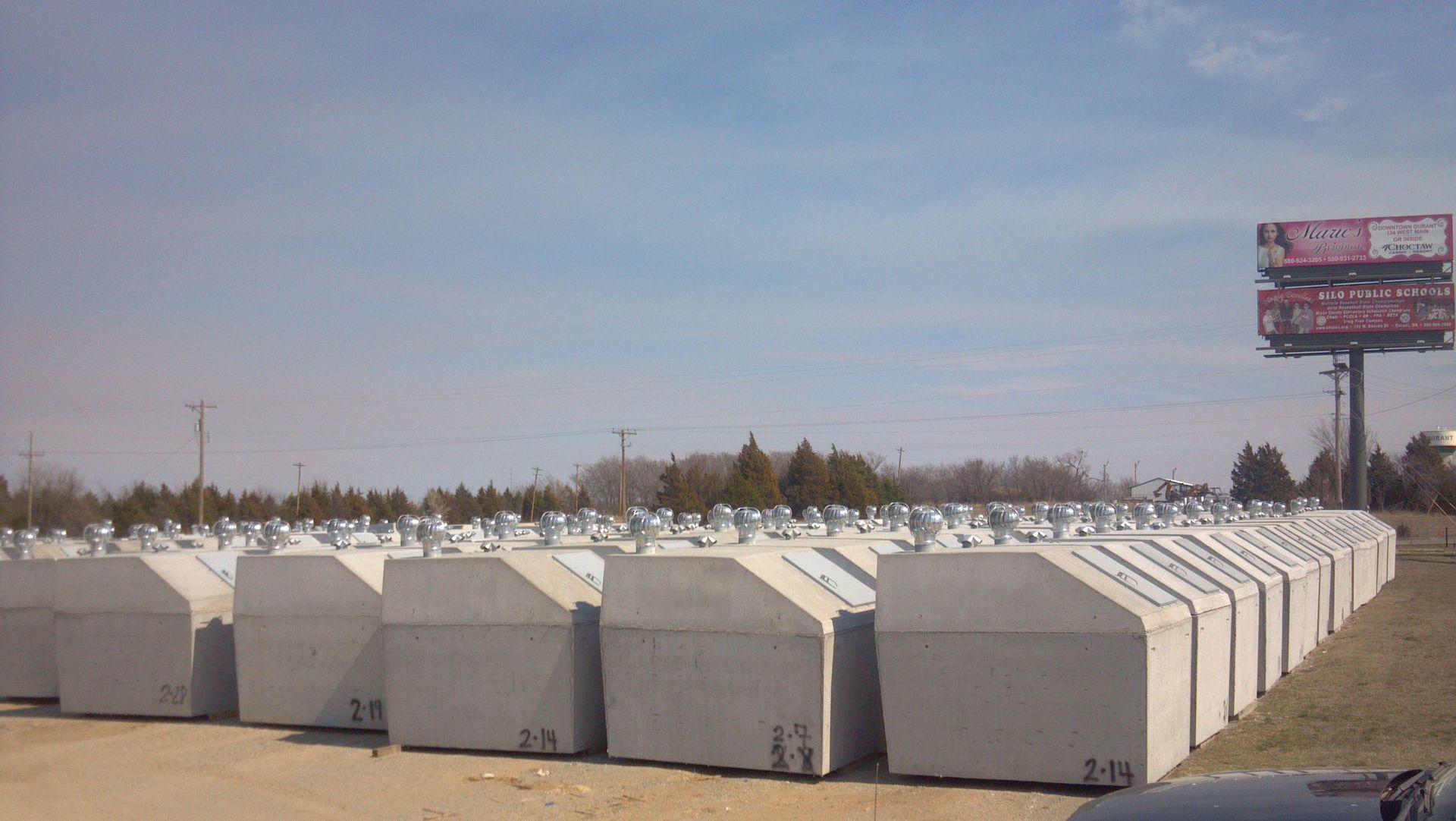 Rows of large, gray concrete structures with metal tops, outdoors under a sunny sky near a billboard.