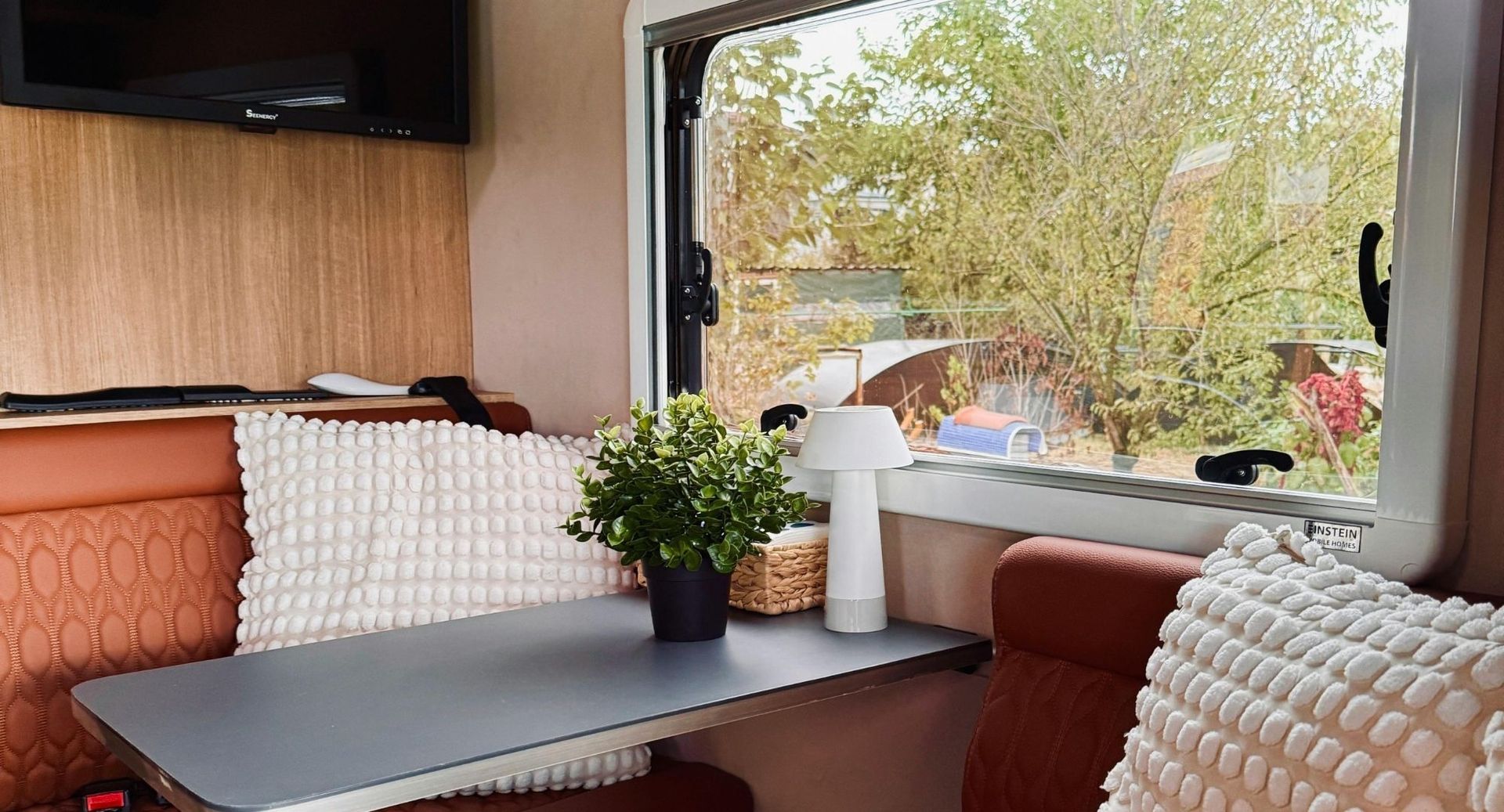 Cozy camper lounge with orange and white cushions, a small table, and a window view of trees and a parked car